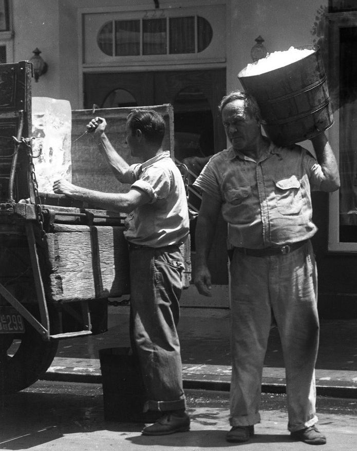 An ice truck is parked as one iceman chips at a block with a pick, and another holds a bucket full of cubes.