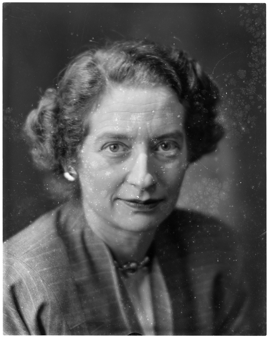 Black and white portrait of an older person with curly hair, wearing a suit and earrings. The image has a vintage, grainy texture. The person is looking slightly to the side with a neutral expression.