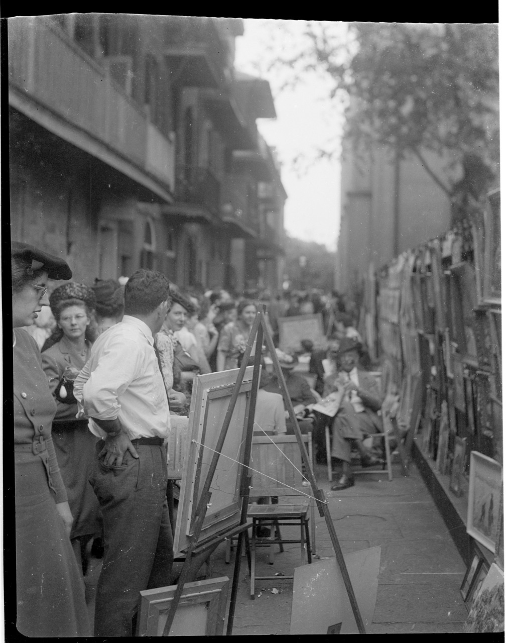 A black and white photo of a bustling street art market. People are browsing paintings displayed on easels and propped against walls. The setting is lively, with many visitors and artists. Buildings and trees line the background.