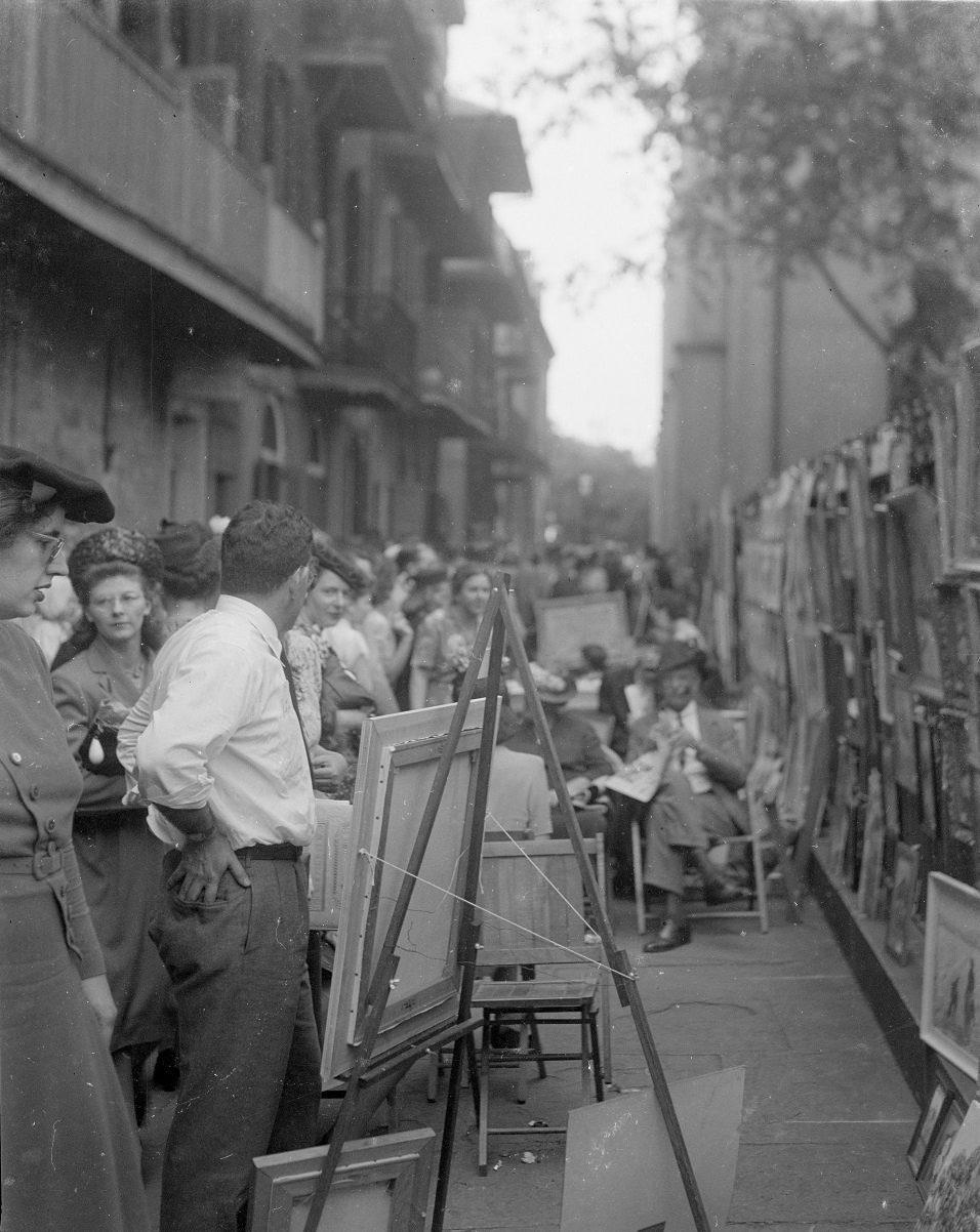 A black and white photo of a busy outdoor art market. People are gathered along a narrow street lined with paintings displayed on easels and against the walls. Several individuals are looking at the artwork while others walk by or observe the scene.