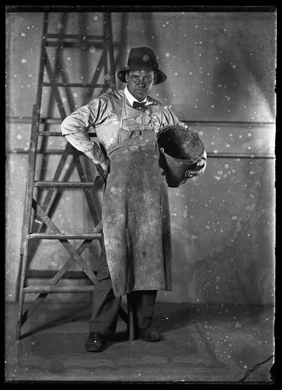 A black and white portrait of a man wearing a hat, glasses, and a striped apron. He holds a welding helmet and stands in front of a wooden ladder against a plain background. The image has visible scratches and marks.