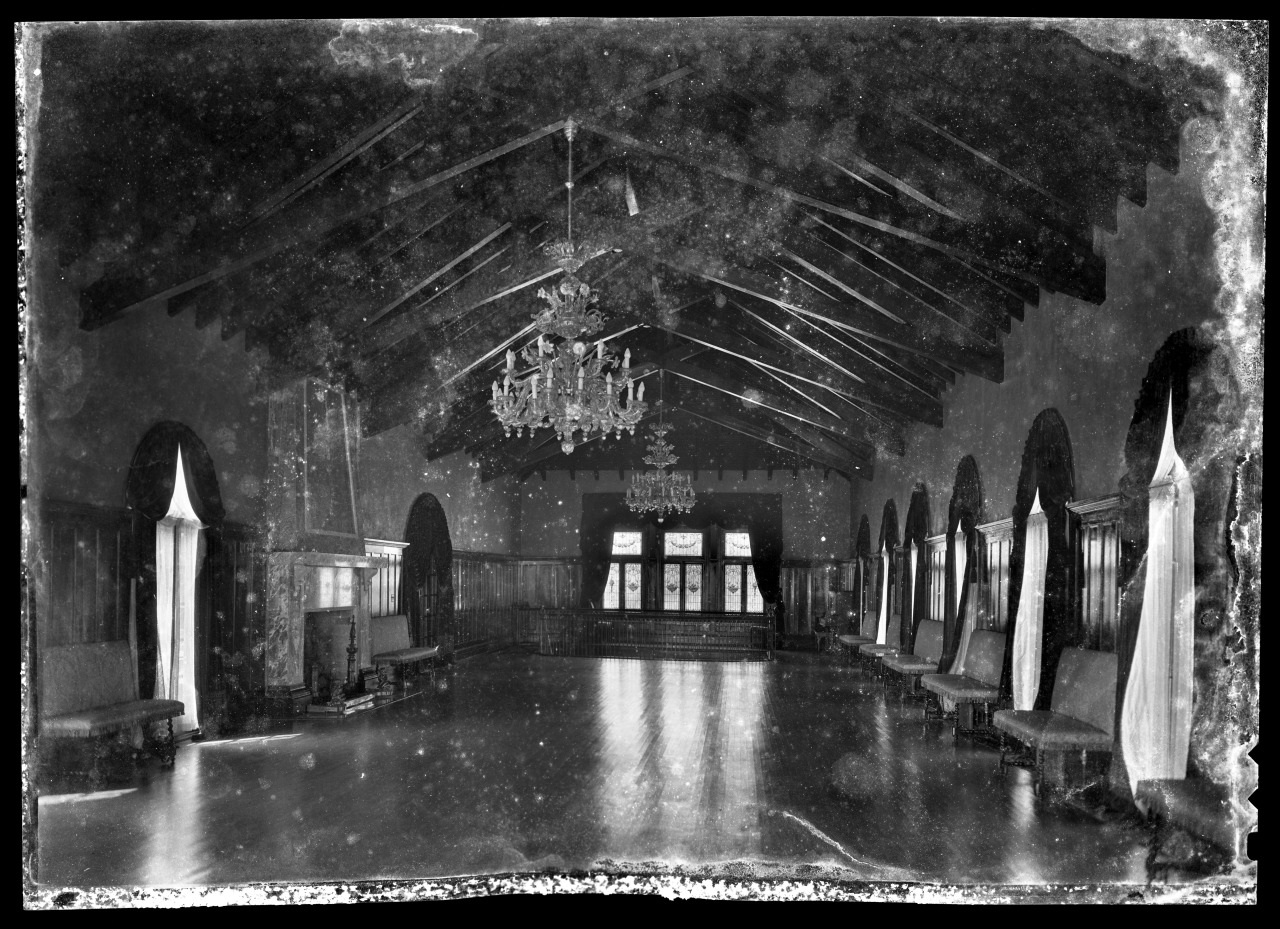 A vintage black and white photo of an ornate ballroom with wooden beams, chandeliers, large windows with draped curtains, and wooden benches lining the walls. The image has a faded, aged appearance.