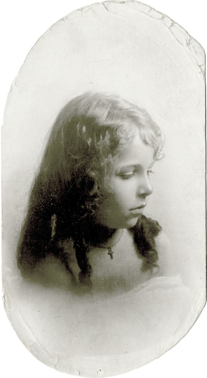 A vintage portrait of a young girl with long, curly hair, looking down. She wears a cross necklace and a dark dress, with a serene expression. The image has an oval border and a soft, faded appearance, giving it an old-fashioned feel.