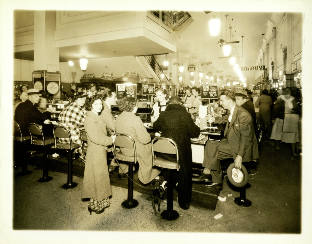People are sitting at a long counter in a busy diner or lunch counter, with a dog underneath. The space is bustling with activity, displaying early to mid-20th century attire and decor.
