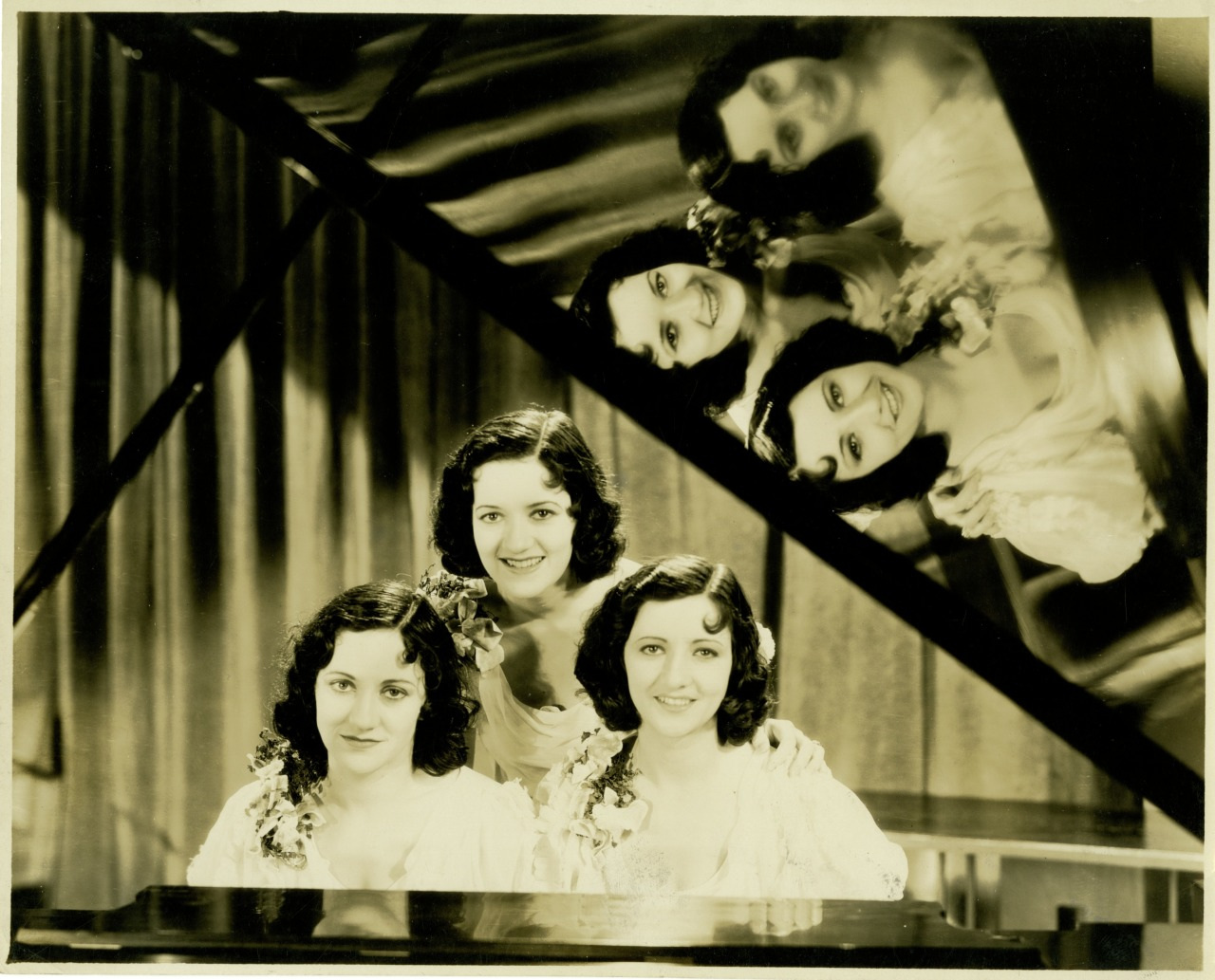 Three women with wavy hair sit closely at a piano, smiling for the camera. Their reflections are visible in the pianos glossy surface, creating a mirrored effect. They are dressed in elegant attire with floral details, and a curtain backdrop is behind them.