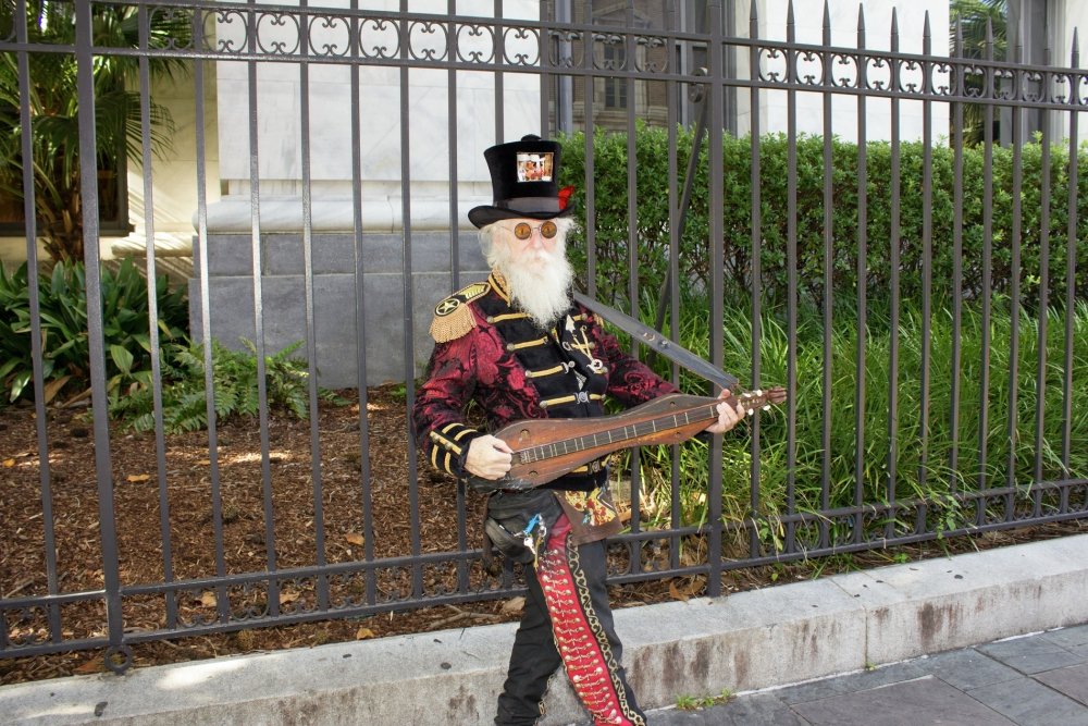 A person dressed in an ornate costume with a top hat and sunglasses plays a stringed instrument in front of a wrought iron fence. Lush greenery and a building are visible in the background.