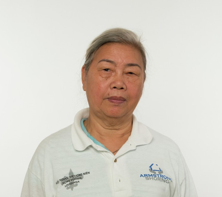 A woman with gray hair wearing a white polo shirt stands against a plain white background. The shirt has logos and text in different languages on it. She is looking directly at the camera with a neutral expression.