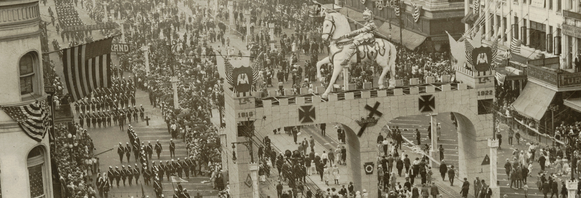A black-and-white image of a busy street parade. Crowds line the sidewalks, and a large archway with a horse statue on top spans the street. Marching bands and numerous people walk in the parade. Buildings with flags and banners flank the scene.