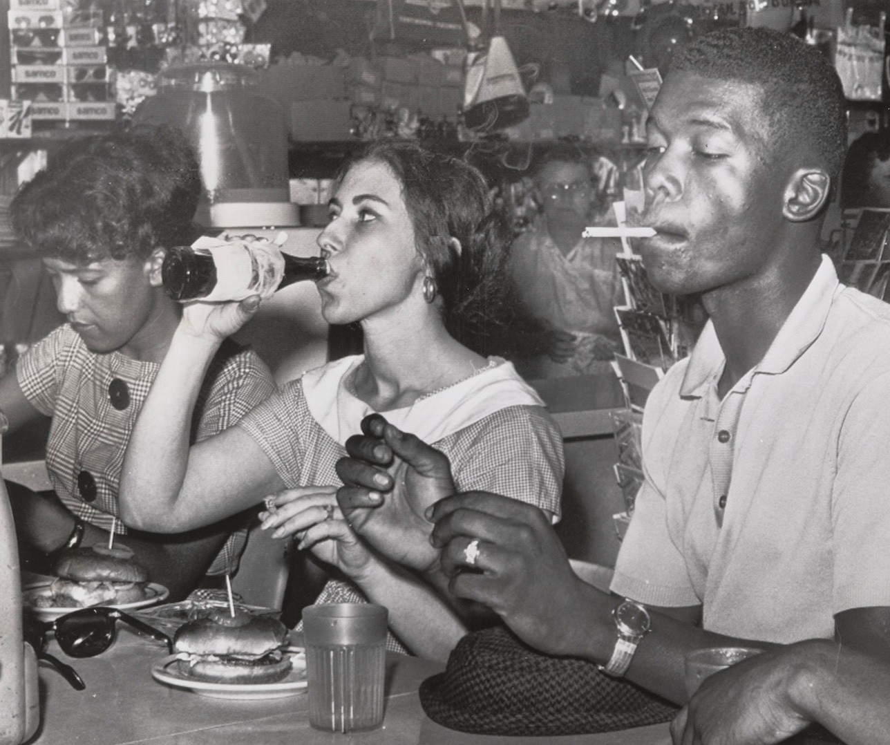 A group of people sits at a diner counter. A woman drinks from a soda bottle, while another eats a burger. A man is smoking a cigarette. The counter is filled with plates and glasses, and the background shows shelves with various items.