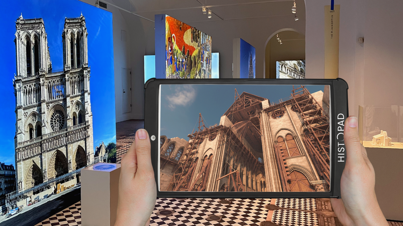 A person holds a tablet displaying a historic image of Notre-Dame Cathedral under construction. The background features modern displays of Notre-Dames architecture and artwork in a museum-like setting.