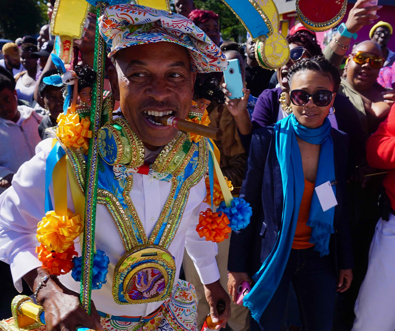 A person in colorful, ornate traditional clothing and hat walks joyfully with a cigar in their mouth. They are surrounded by a crowd of people, including a person in sunglasses and a blue scarf. The scene is lively and festive.