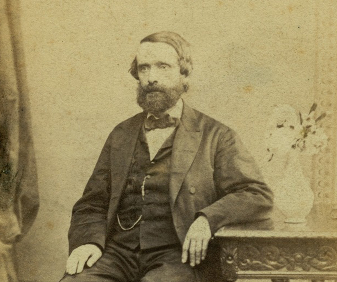 A sepia-toned vintage portrait of a bearded man sitting beside an ornately carved table with a decorative vase. He wears a three-piece suit and has a calm expression, with his hand resting on the table.