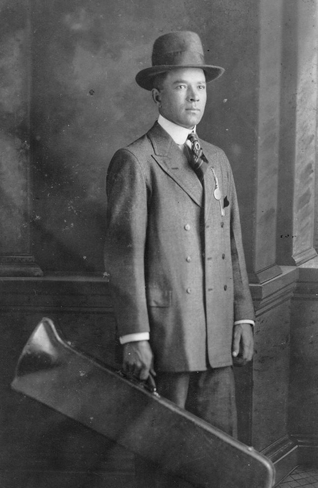 A vintage photograph of Kid Ory, taken in 1917. He wears a dark suit and hat and carries a trombone case.