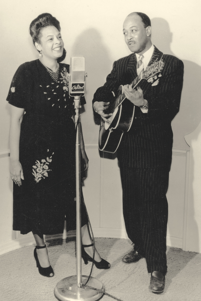 A woman singing into a vintage microphone, wearing a dark dress with floral embroidery. Beside her, a man in a dark pinstripe suit is playing an acoustic guitar. They appear to be performing together in a studio.
