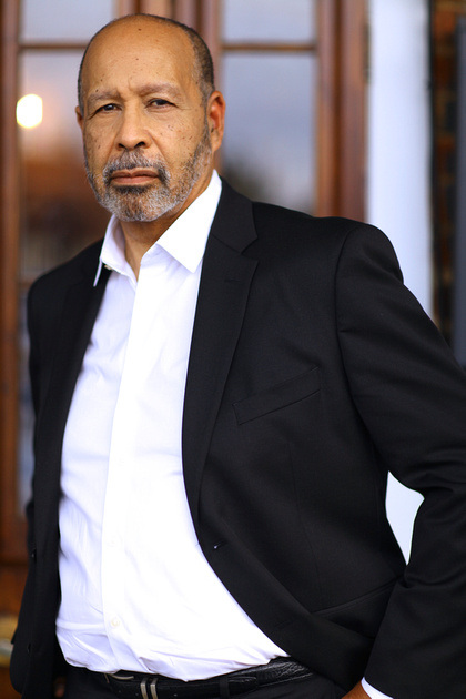 A man with a beard wearing a black suit and white shirt stands with one hand in his pocket, looking directly at the camera. The background shows a wooden door and a blurry exterior scene.