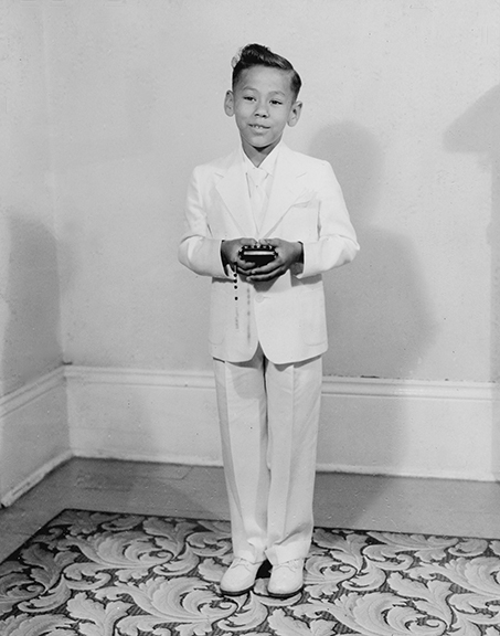 A vintage black and white photo shows Larry on the day of his first Communion as a young boy in 1954. He is smiling while wearing an all-white suit.