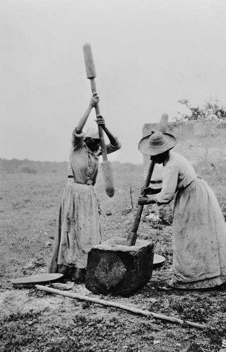Two women in historical attire use large wooden pestles to pound food in a stone mortar outdoors. They are wearing long skirts, aprons, and straw hats, working together in a rural setting.