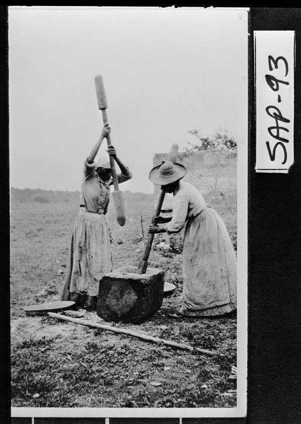 Two women in long dresses and hats are using large wooden tools to pound in a rural setting. They are standing beside a large stone block, working together. The photo is in black and white and labeled SAP-93 on the side.