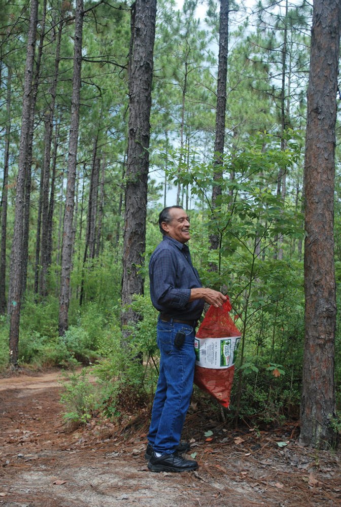 A man in jeans and a dark shirt stands in a forest, holding a red bag. He is smiling and surrounded by tall pine trees and greenery.