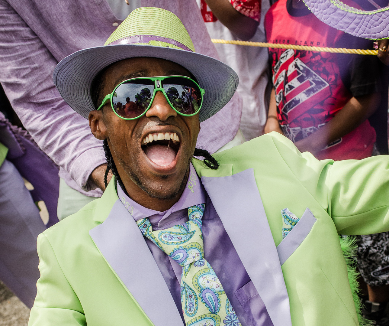 A joyful person wearing a lime green and lavender suit, matching hat, and reflective green sunglasses is smiling widely. They are surrounded by others, with part of a red and white shirt visible in the background.