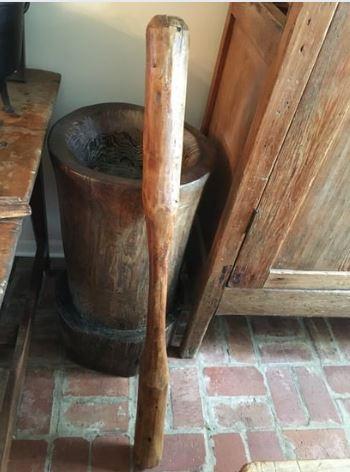 A wooden pestle leans against an old wooden mortar on a brick floor. The scene is in a rustic setting with wooden furniture.