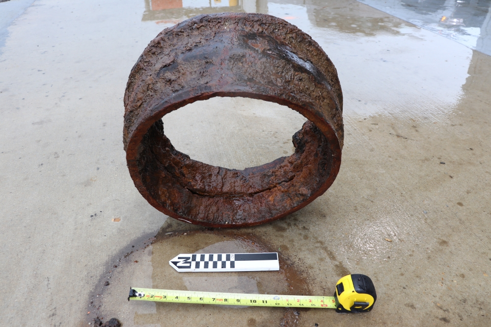 A rusty, corroded metal pipe section lies on wet concrete. Beside it are a measuring tape and a scale bar for reference. The measuring tape shows the pipes diameter, and the scene suggests recent rain or wet conditions.