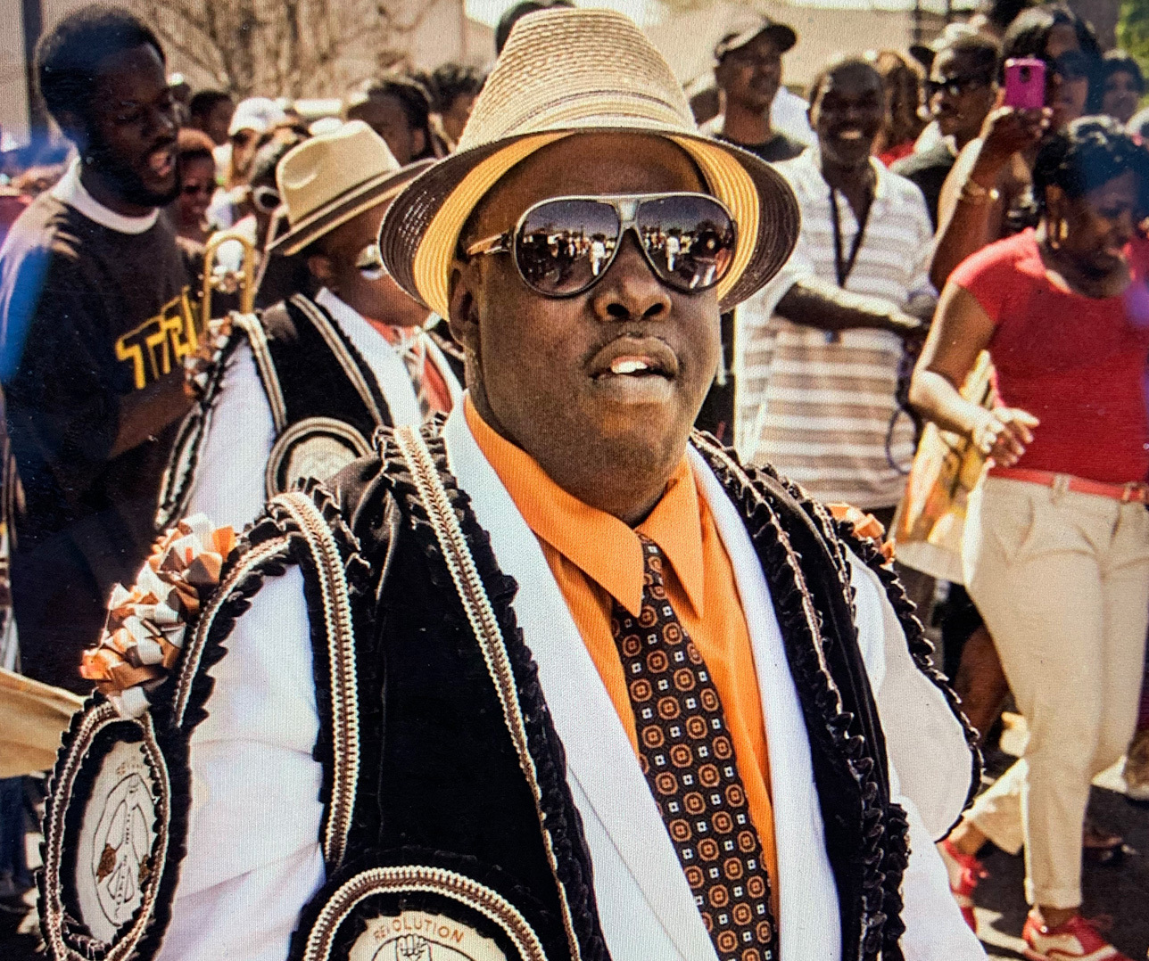 A man wearing sunglasses, a straw hat, and a vibrant orange shirt with a patterned tie participates in a lively parade. He is surrounded by a crowd of onlookers and other parade participants in decorative attire.