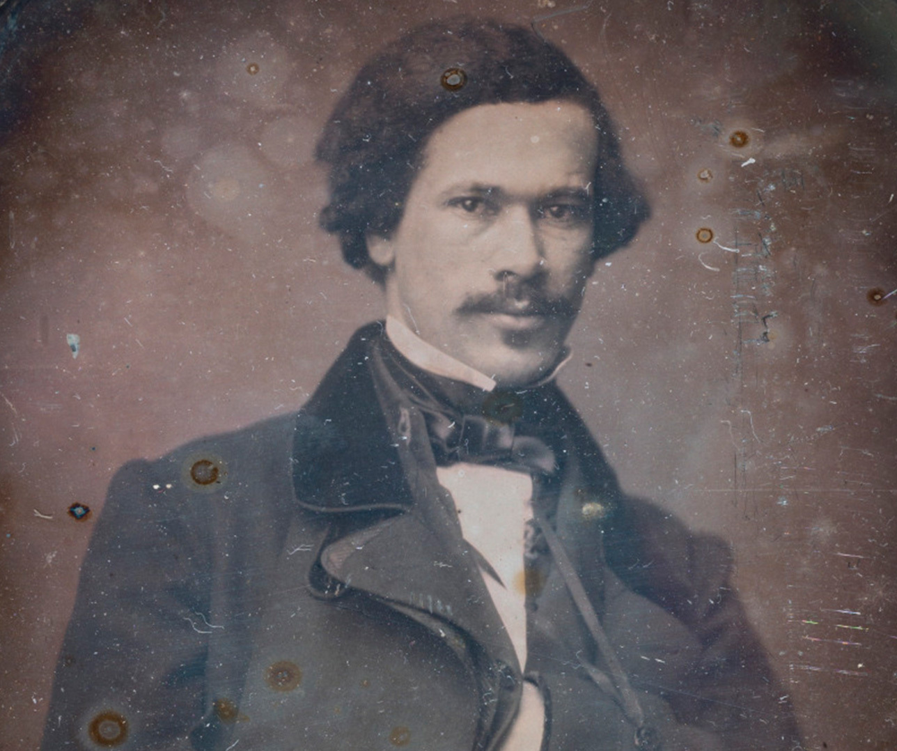 A sepia-toned vintage photograph of a man with curly dark hair and a mustache. He is dressed in a 19th-century suit with a double-breasted coat and bow tie. The image has an old, slightly worn appearance.