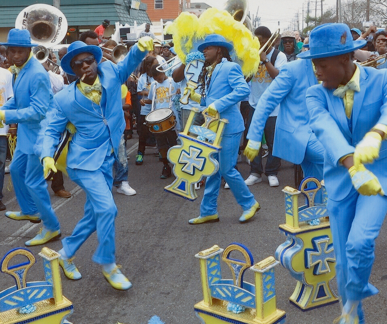 A vibrant street parade features performers in bright blue suits and hats, energetically dancing. Theyre surrounded by musicians and colorful decorations, with a lively crowd enjoying the festive atmosphere.