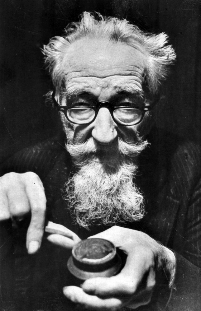 An elderly man with glasses and a long white beard holds a magnifying glass and examines a small object. He has unruly hair, a focused expression, and is dressed in a pinstripe suit. The image is in black and white.