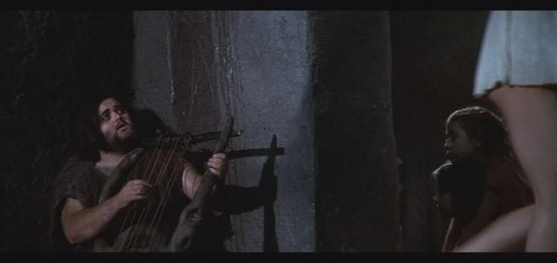 A man sits against a wall playing a stringed instrument, possibly a lyre. Two onlookers, partially visible, watch him intently. The scene is dimly lit, suggesting a historical or ancient setting.