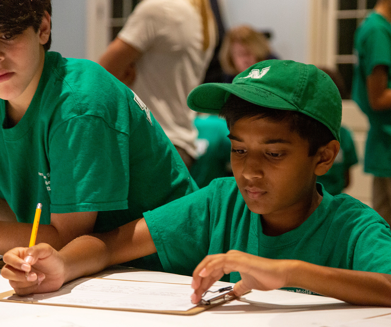 A young boy in a green shirt and cap writes on a clipboard with a pencil. He is focused, and another person wearing a similar shirt is partially visible beside him. The background shows more people and a window.