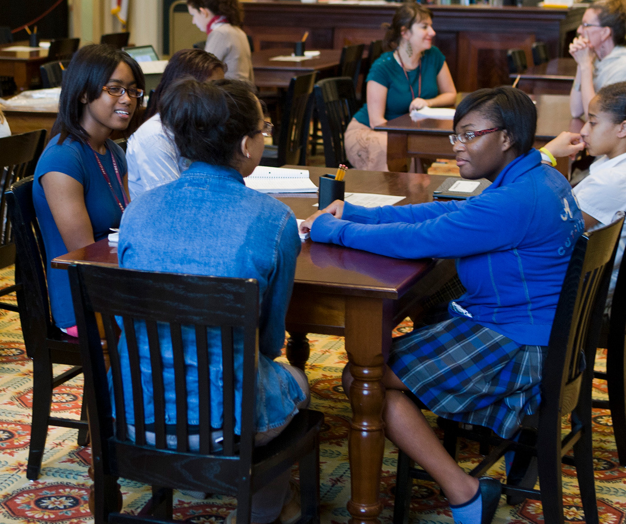 A group of students sitting around a table in a classroom, engaged in discussion. The classroom has wooden furniture and a patterned carpet. Other students and a teacher can be seen in the background, listening and participating.