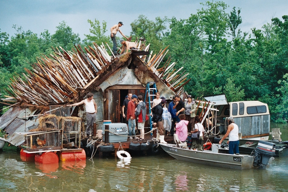 A group of people work together on a floating, makeshift house with wooden spikes on the roof. Some stand on the roof, while others are in a small boat alongside. The scene is set on a river, surrounded by lush greenery.