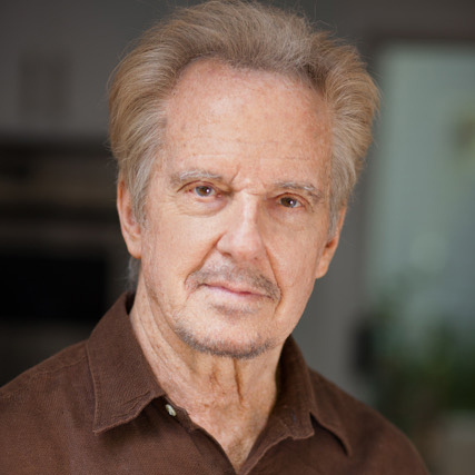 Portrait of an older man with short, light hair and a light beard. He is wearing a brown shirt and looking directly at the camera. The background is softly blurred, emphasizing his face.