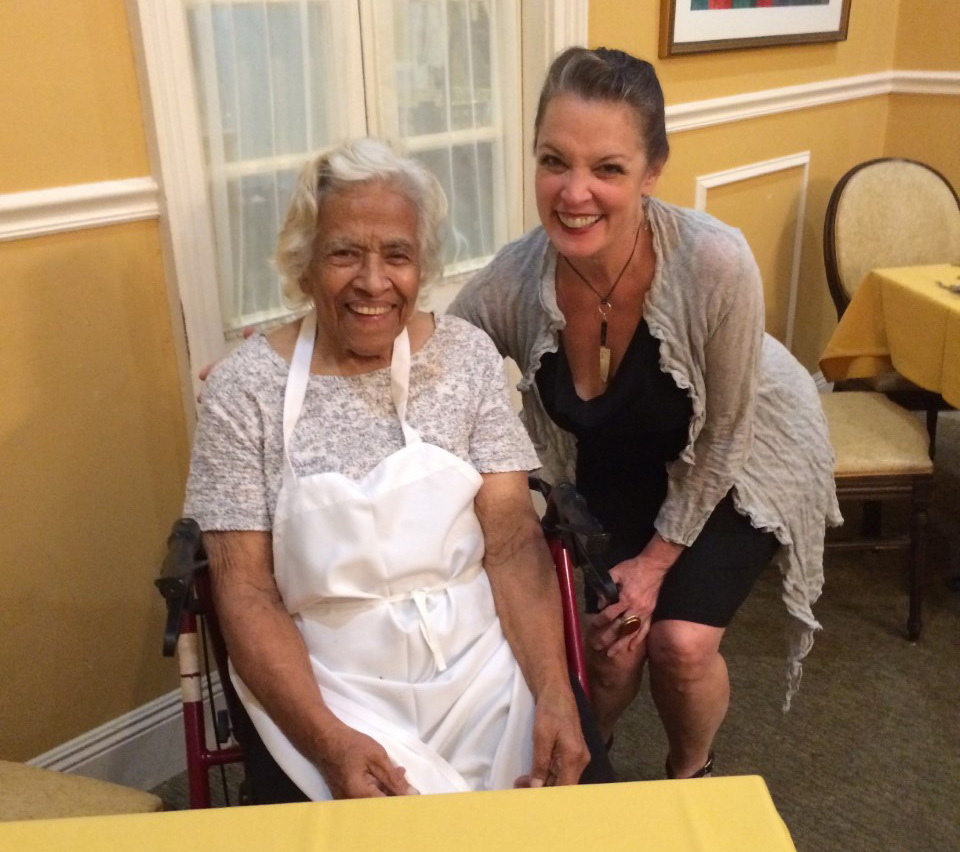 An elderly woman with white hair smiles while sitting in a wheelchair wearing a white apron. A middle-aged woman with brown hair in a black dress and gray shawl leans beside her, smiling. They are indoors with yellow walls and a dining room setting.