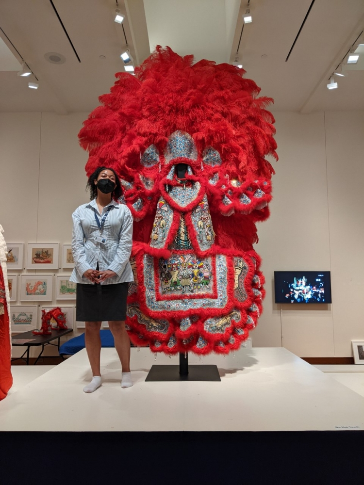 A person stands next to an elaborate, red feathered costume adorned with intricate beadwork and designs on display in a museum. The person is wearing a mask and a light-colored outfit. The background features framed artwork on the walls.
