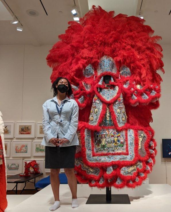 A woman stands next to an elaborate costume made of red feathers with intricate beadwork. Shes wearing a mask, a striped blouse, and a black skirt. The display is in an art gallery or museum setting.