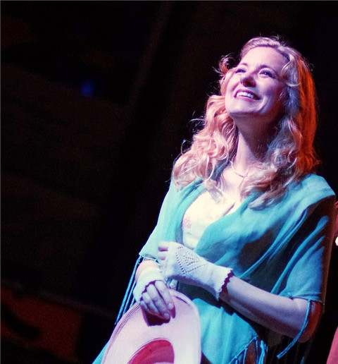 A woman with curly blond hair stands on stage, smiling and looking upward. She holds a hat and wears a turquoise shawl over a light dress. The lighting highlights her joyful expression against a dark background.