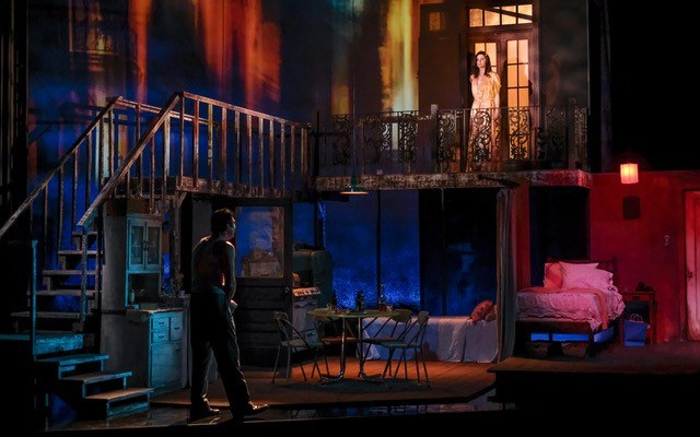 A theatrical stage scene with a dimly lit, vintage apartment set. A woman stands on an upper balcony, looking down at a man in a dark tank top on the lower level. The room includes a bed, table, chairs, and a staircase.