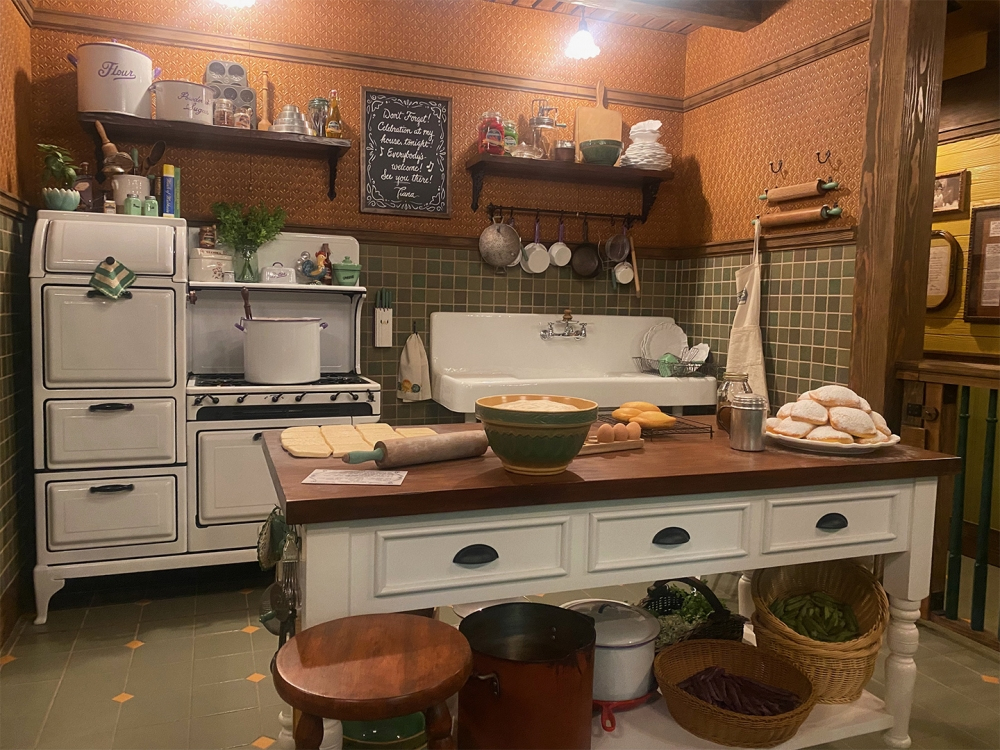 A vintage kitchen with a white stove, a large sink, and a wooden island. There are various kitchenware items, vegetables, and baked goods on the island. Shelves with jars and decor line the wall above the sink, and a chalkboard hangs nearby.