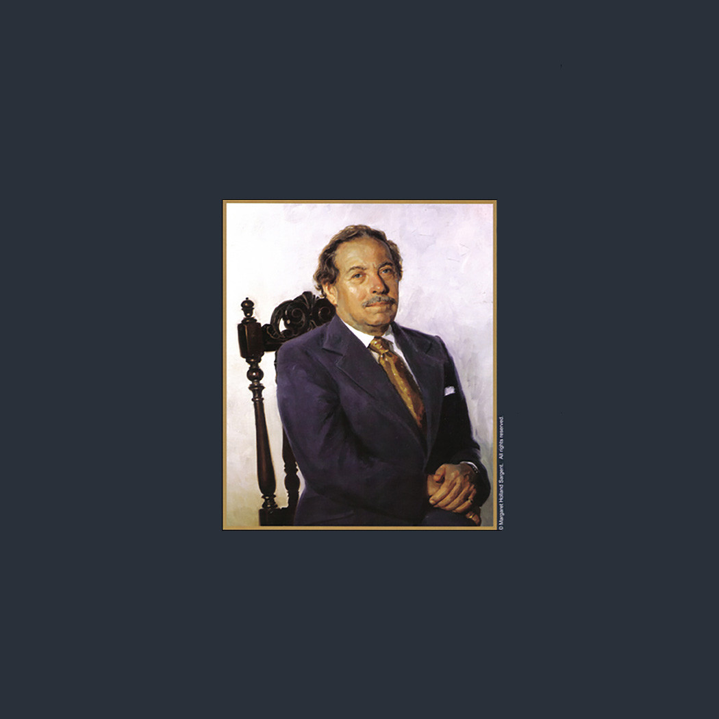 A seated man in a dark suit and tie poses for a portrait against a light background. He sits on an ornate chair, his hands folded neatly in his lap, exuding confidence and calmness.