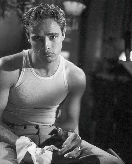 A black and white photo of a man wearing a sleeveless shirt and high-waisted trousers, sitting with a serious expression. He holds a piece of cloth in one hand. The setting appears to be dimly lit and indoors.