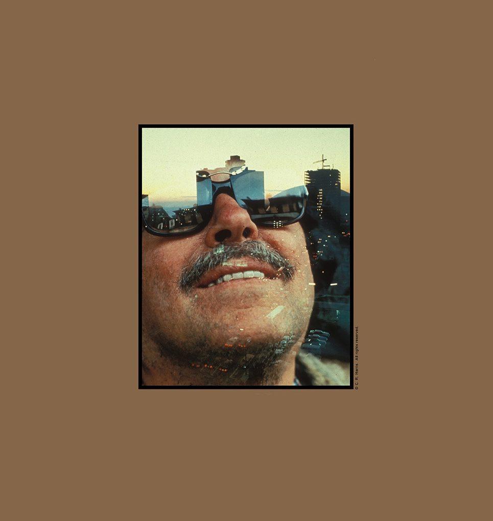 A close-up of a man wearing reflective sunglasses, with a cityscape mirrored in the lenses. He has a mustache and is smiling slightly. The background is a brown frame around the image.