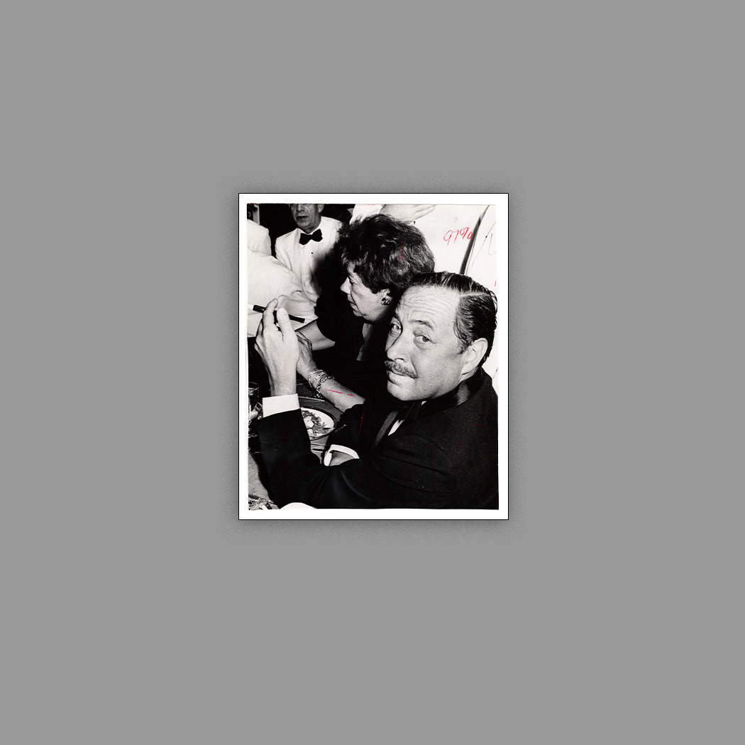 A black and white photo of a man in a suit looking at the camera while seated at a dining table. A woman is next to him, leaning over, facing away. Other diners in formal attire are visible in the background.