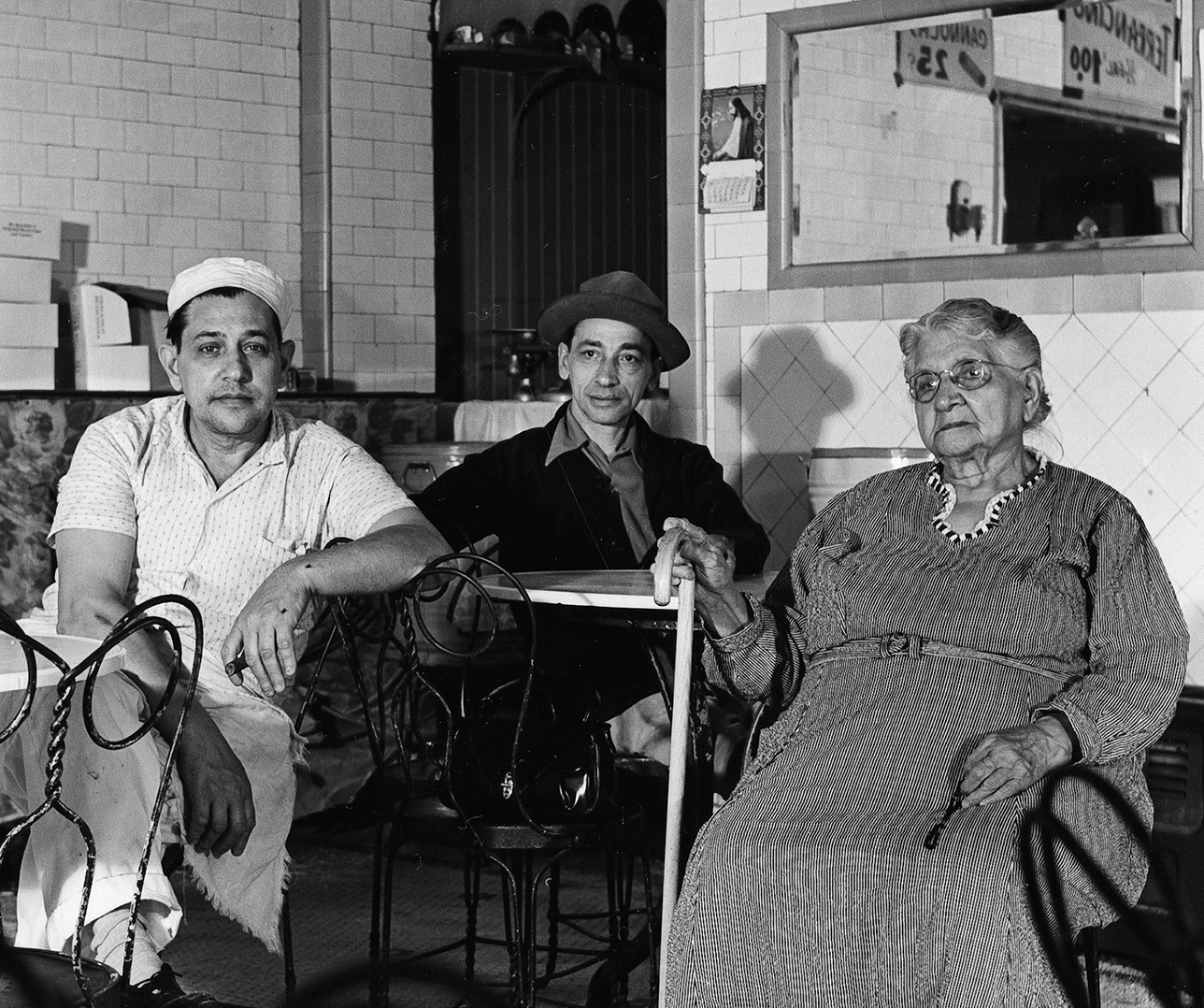 Three people sit inside a diner. From left to right: a man in a white shirt and cap, a man in a dark jacket and hat, and an elderly woman in a dress with a cane. They are seated around a small table with chairs. The setting is cozy and vintage.