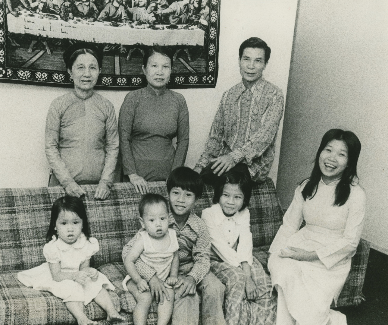 A vintage family photo of nine people. Four adults stand behind a sofa with five children sitting on it. The background features a tapestry. Everyone is smiling, capturing a moment of joy and togetherness.