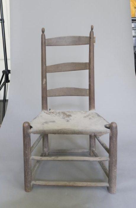 A worn-out wooden chair with a high backrest and ladder-style design. The seat is frayed and damaged, revealing the underlying material. The chair sits on a plain gray background.