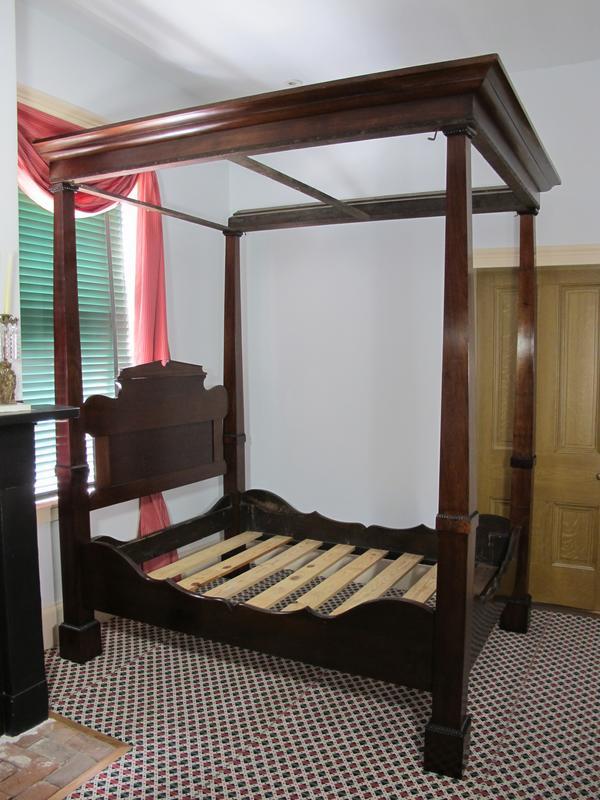 A large wooden four-poster bed frame with no mattress stands on a patterned carpet. It features a headboard, footboard, and canopy structure. A red curtain hangs on a window to the left, and a wooden door is visible to the right.