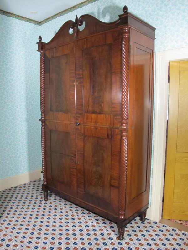 A large, ornate wooden wardrobe with intricate carved details stands against a patterned wallpaper background. The wardrobe features detailed columns on its sides and a curved top. The room has a patterned carpet and a visible door to the right.